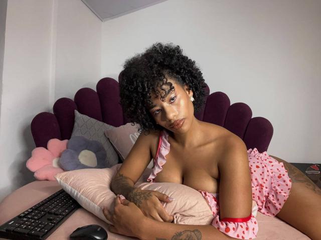 KarlaRicochet, con cabello negro rizado, piel morena y tatuajes, está acostada en una cama con ropa interior rosa de lunares, cubriendo sus pequeños senos con un cojín rosa..
