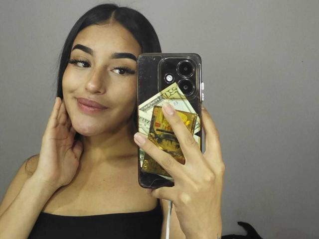 ElowenGrace with black hair, wearing a black top, holding a phone with a money clip case, smiling, hands near face, grey background. Phone case has visible dollar bills.