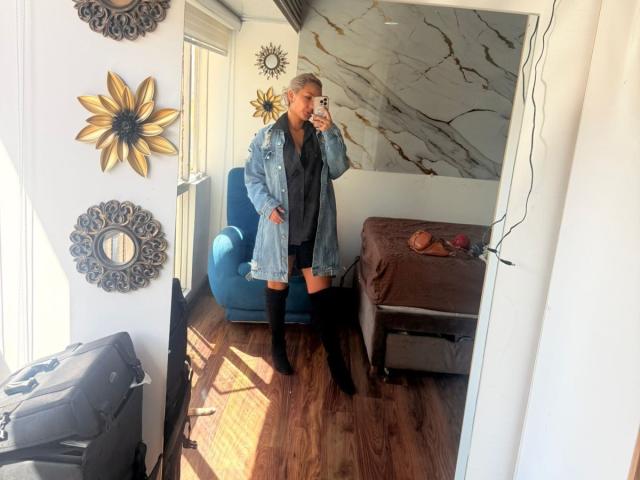 ElenaSaenz with blonde hair, wearing a blue denim jacket and black thigh-high boots, taking a mirror selfie, showing her ass in a dark dress, in a sunlit room.
