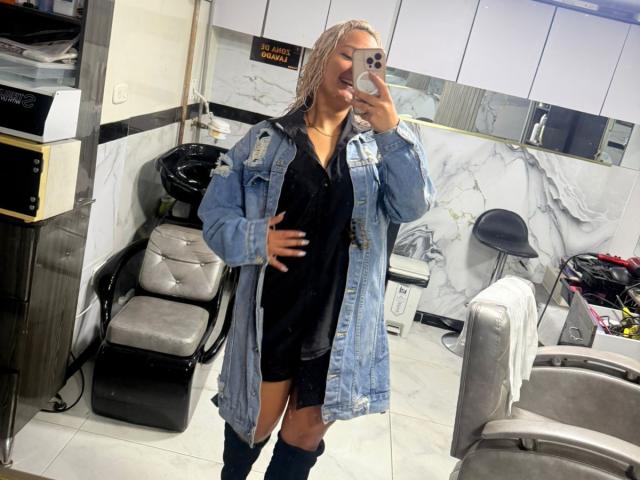 ElenaSaenz in a blue denim jacket over a black dress, standing in a salon, taking a mirror selfie with a phone, smiling, blonde hair, knee-high boots.