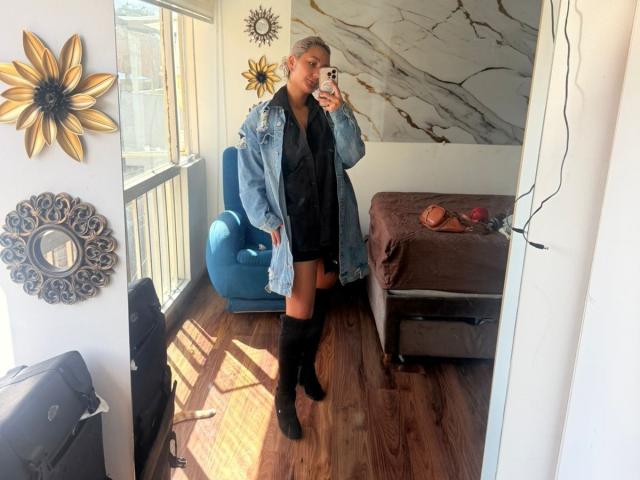 ElenaSaenz with short blonde hair, wearing a denim jacket, black shirt, and thigh-high boots, taking a mirror selfie in a sunlit room. Boobs not visible, ass not visible.