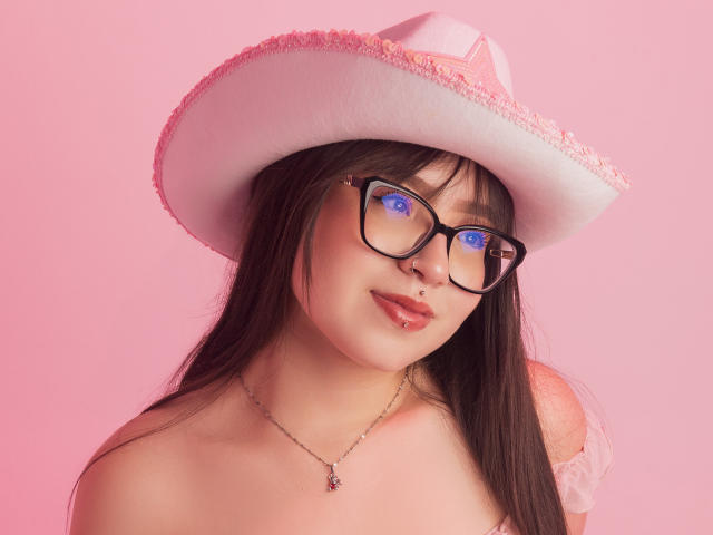 MadeleynRouse with dark hair, glasses, pink cowboy hat, nose piercing, lip piercing, necklace, bare shoulders, pink background, blue-eyed reflection in glasses.