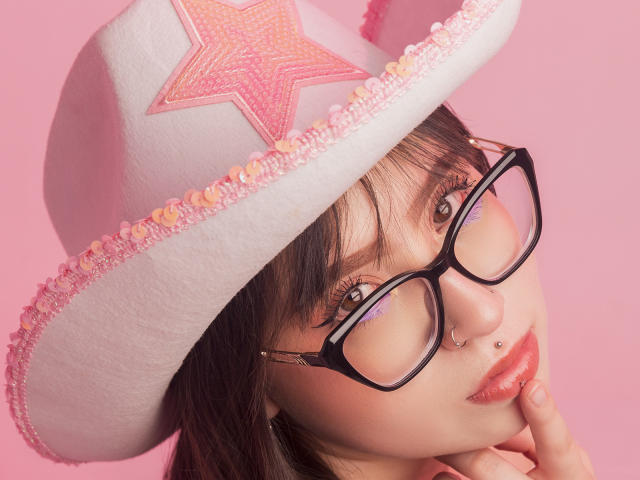 MadeleynRouse with glasses, white cowboy hat with pink star and beads, pink background, nose ring, and finger near lip, looking up, showing cute and sexy.