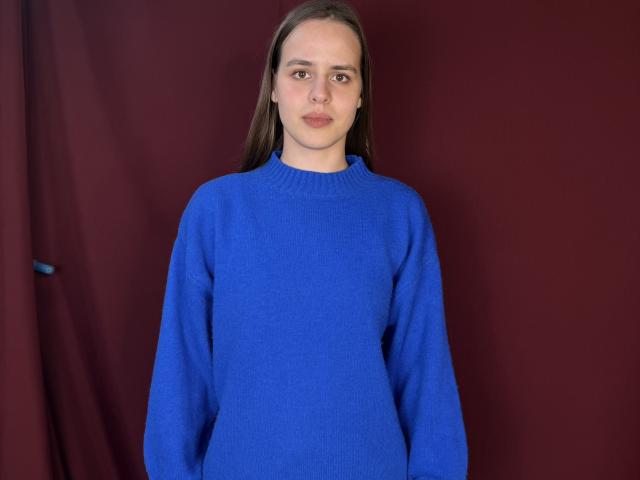 SandiWimbs, com cabelo castanho liso, vestindo um suéter azul folgado, em pé contra um fundo vermelho escuro, olhando diretamente à frente com uma expressão neutra..