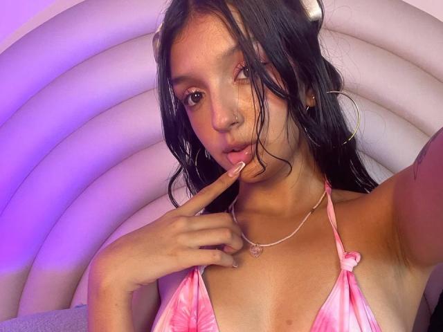 PaulineRey met lang zwart haar, een roze bikini top, een vinger op haar lippen, een schattige neusring, gouden oorringen, een roze ketting, paars-blauw licht en een zachte, witte, gebogen achtergrond..