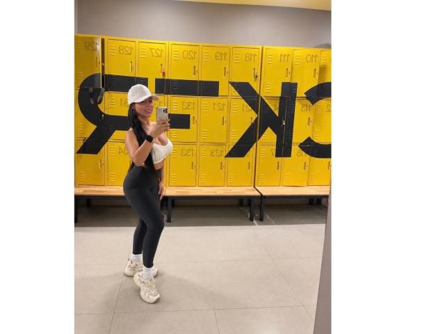 NahomyStone in a white cap, sleeveless top, black leggings, and white sneakers, taking a mirror selfie in a gym locker room with yellow lockers.