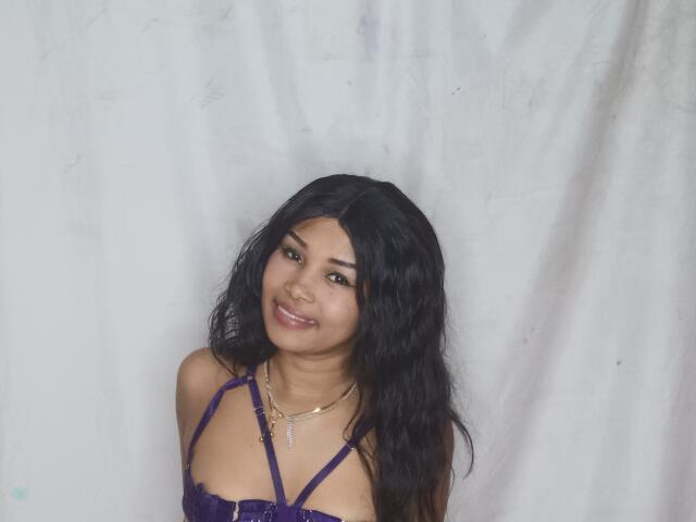 LaCoquineX with long black wavy hair, wearing a purple lace bra, smiling at the camera, has medium brown skin, and a gold necklace with a small cross.