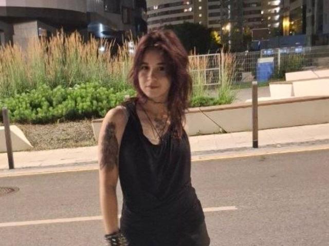 ShenikaTrucker with wavy dark red hair, black sleeveless dress, tattoos on her right arm, standing on a city street at night, smiling slightly, looking relaxed.
