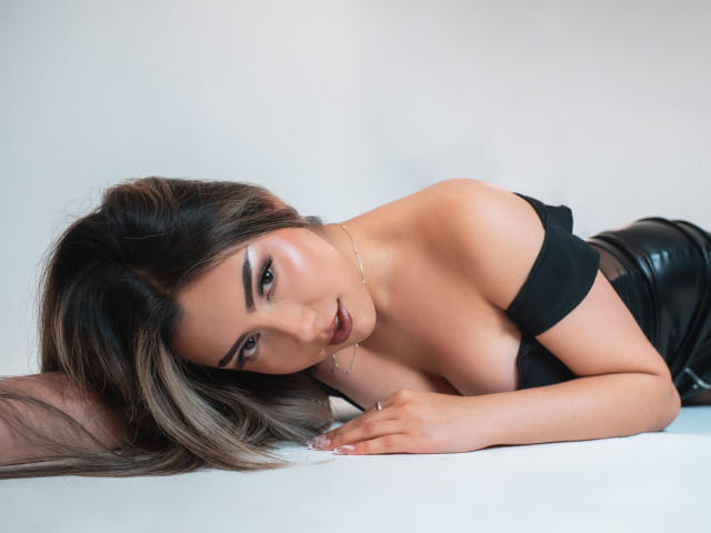GabrielaMonique with long brown hair, lying on white, black strap dress, showing her tits, black leather pants, gold necklace, seductive gaze, parted lips, and soft makeup.