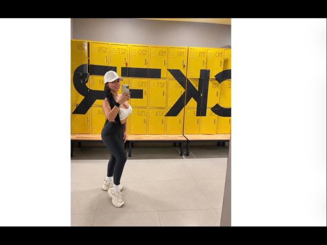 NahomyStone with white hat, white top, black pants, and white sneakers, standing in front of yellow lockers, holding phone, showing curvy ass and small tits, in gym locker room.