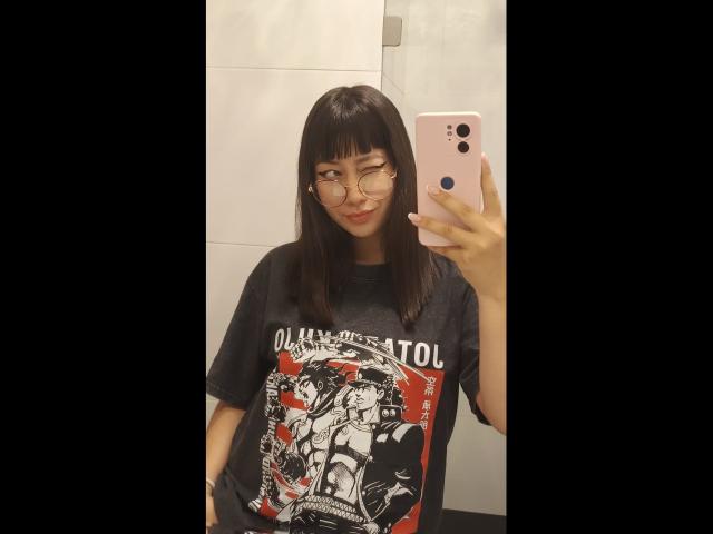 Genesiss with straight black hair, glasses, and a gray Ozzfest t-shirt, holding a pink phone, smiling, showing off her small tits in a bathroom mirror selfie.