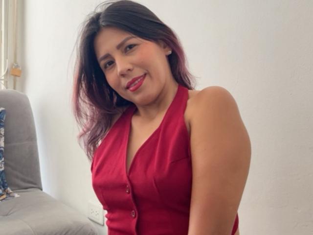 LunaTomson with medium tits, wearing a red button-up sleeveless top, has dark brown hair with purple tips, and a cute smile with red lipstick, in a casual setting.