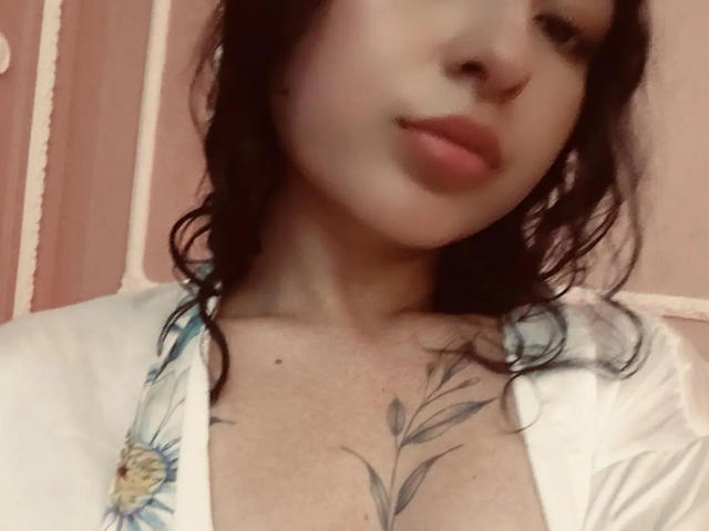 MiiaBennet with wet black hair, slightly parted lips, white floral shirt, and visible tattoo on chest, showing small tits. Her nose is pierced. Soft, pink background.