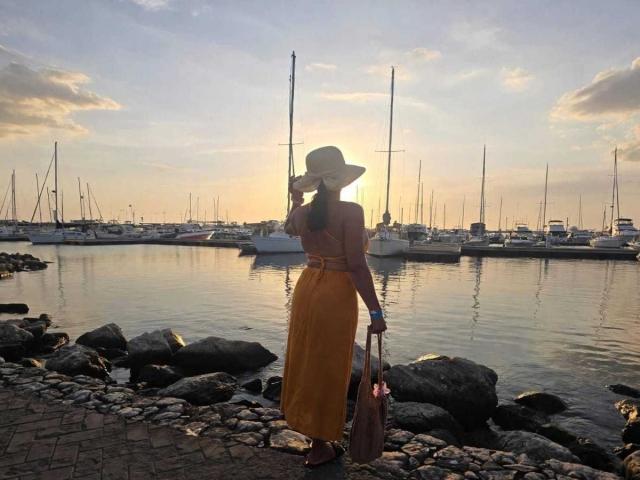 JadeFoster stands on a rocky waterfront, back to the camera, in a yellow dress and hat, holding a bag, sun setting, sailboats in calm water, clouds in sky.