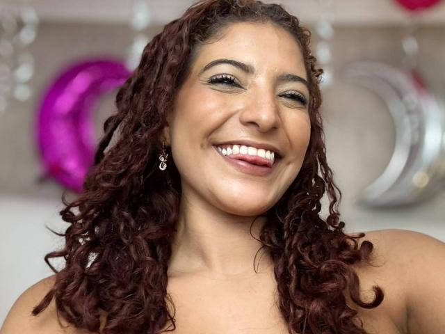 MiaBianchi has curly brown hair, tan skin, and is smiling with her tongue slightly out. She's wearing earrings and has white teeth. Boobs are not visible.