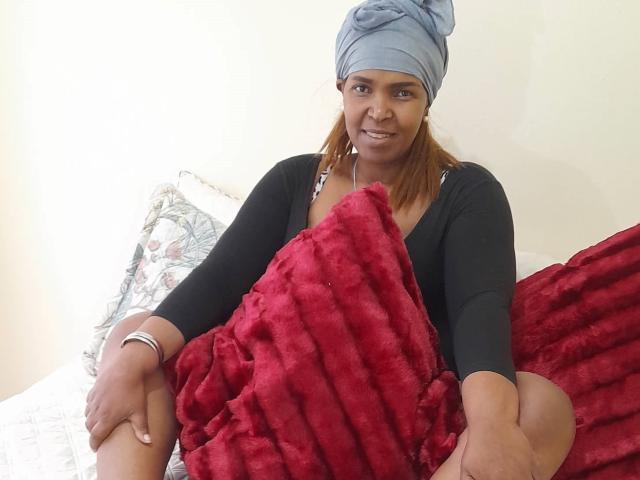 MeganBrownskin with brown skin, wearing a blue headwrap and black top, sitting on a bed with red, fluffy butt in the foreground. She's smiling and holding the butt.
