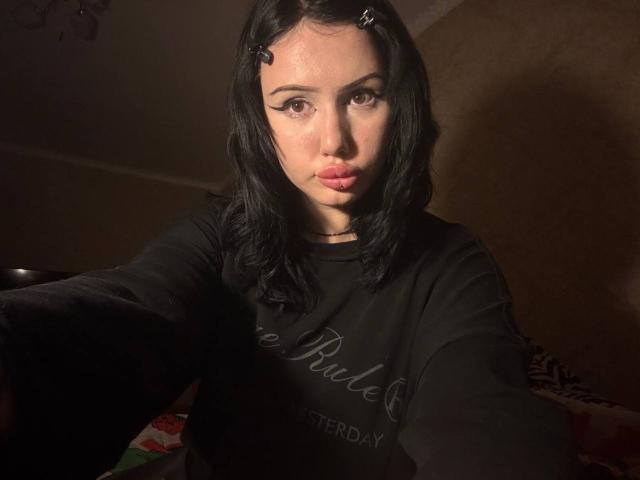 SundayScholzen, con i capelli scuri, indossa una maglietta nera, ha labbra rosa carnose e sta fissando la fotocamera con un'espressione leggermente seria, illuminata lateralmente..