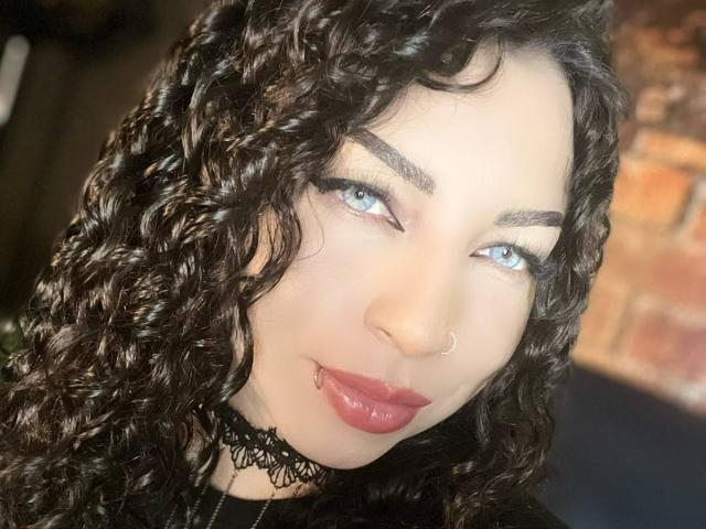ScarlattaaDesire has curly black hair, blue eyes, a nose ring, and red lipstick with a lip piercing. She's wearing a black lace choker. Her tits are not visible.