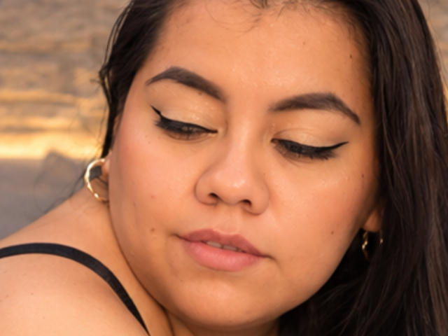 ChubbyThickMilf has tan skin, dark eyebrows, and black eyeliner, with her eyes closed, wearing gold hoop earrings, and a black strap. Her lips are slightly parted.