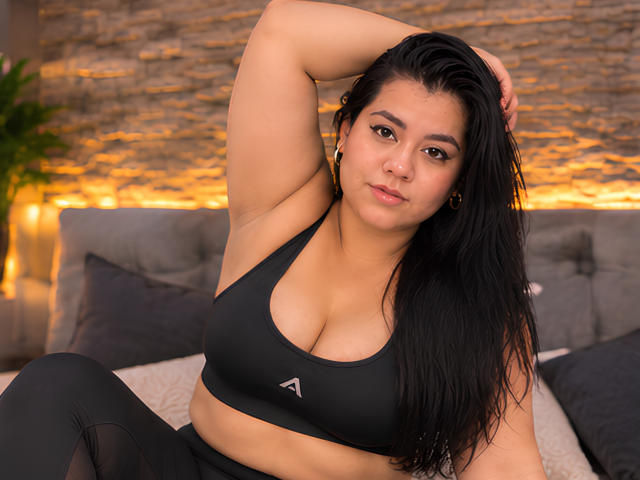 ChubbyThickMilf with black hair, tan skin, and large boobs in a black sports bra, sitting on a bed, arm raised, looking at the camera, wearing black pants.