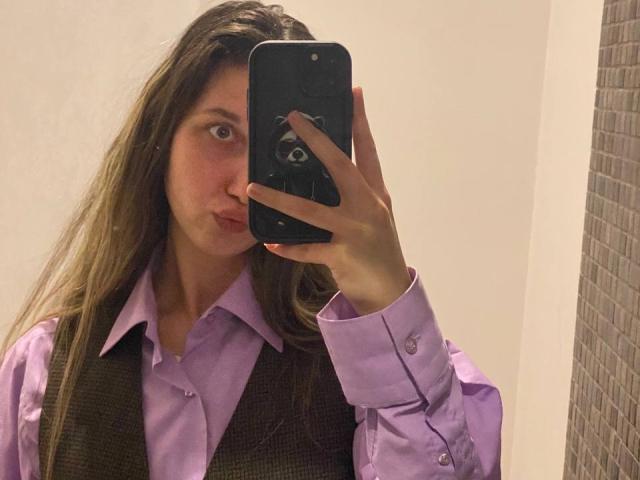 Sanella with long brown hair, puffy pink lips, wearing a purple shirt and brown vest, taking a mirror selfie with a black phone, showing her face and upper body.
