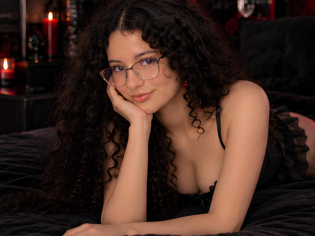 FluffydirtyMind with curly black hair, glasses, and pink lips, resting on black bed, wearing black lingerie, showing small tits, and a cute smile, surrounded by red candles.