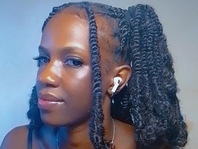 SexyAurora with dark brown skin, glossy lips, and thick black braided hair, wearing white earbuds, looking to the side with a slight smile. Her tits are small.