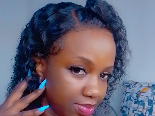SexieAurora, avec ses cheveux noirs bouclés, ses ongles bleus, ses lèvres roses et un piercing au nez, regarde l'appareil photo avec un léger sourire, sa main touchant sa joue..