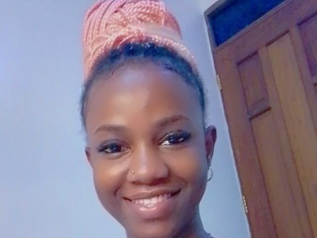SexieAurora with dark brown skin, smiling, wearing a pink knitted headwrap, hoop earrings, and a nose ring, showing teeth and eyes. Wooden door in background.