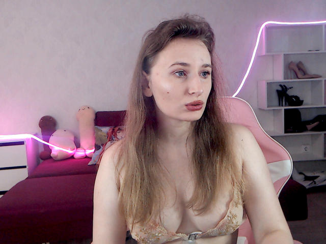 AliceReeve has pale skin, long brown hair, and small tits in a floral bra. She's sitting in a pink chair with a purple neon light behind her.