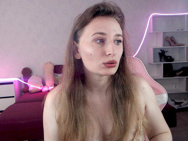 AliceReeve with long brown hair, light skin, pouty lips, small moles on her face, looking to the side, topless, pink neon light in background, white chair behind her.