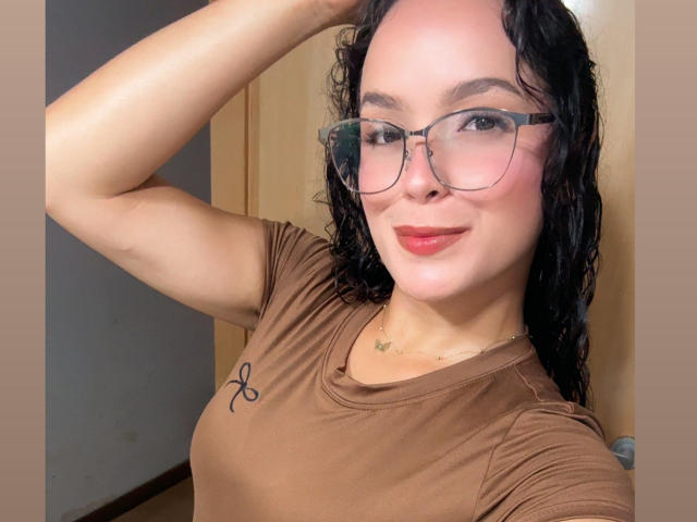 MaiaPark with curly black hair, glasses, brown shirt, small butterfly tattoo on chest, smiling, hand in hair, cute, pink lipstick, medium skin, small breasts, gold necklace.