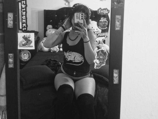 JadeMilleer is in a black tank top, checkered panties, and thigh-high socks, sitting on a bed, taking a mirror selfie, showing her ass and tits. Black and white.