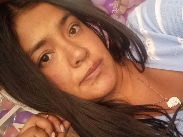 ArianithaXox with long dark hair, brown eyes, and freckles, wearing a blue and white striped shirt, looking at the camera, wearing a silver necklace with a heart pendant.