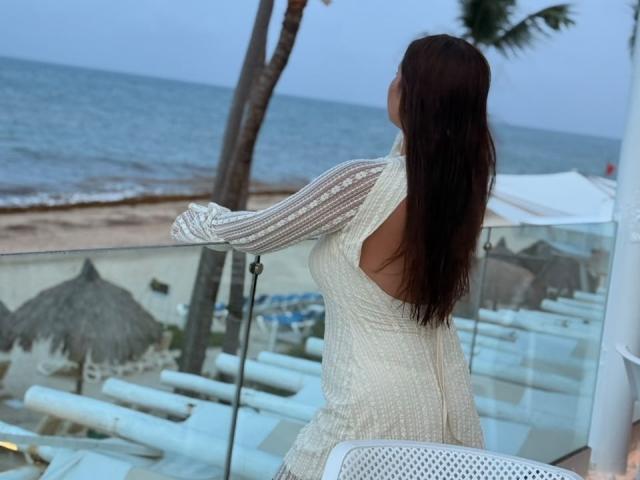 LinaFoxyHot con lunghi capelli castani, di spalle, in un abito bianco trasparente con spalline, appoggiata a una ringhiera di vetro, che mostra il sedere, su un balcone affacciato sulla spiaggia..