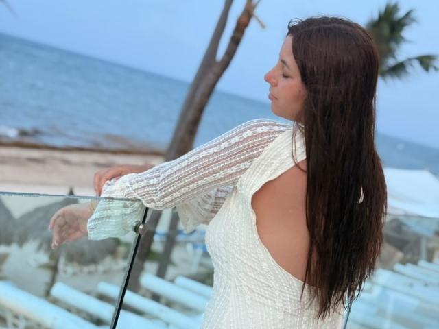 LinaFoxyHot with long brown hair, white open-back dress, and sheer sleeves, leaning on a glass railing, ocean and palm tree in the background, side profile, eyes closed.