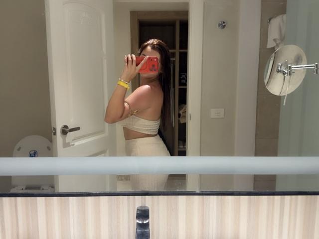 LinaFoxyHot with long brown hair, holding a pink phone, wearing a white strapless top and high-waisted pants, taking a mirror selfie, showing her ass and a yellow bracelet.
