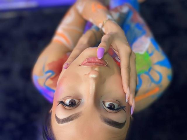 LunaixMoon, upside-down, with painted face, purple nail, ring nose, colorful tattoos, and blue eyes, touching nose with pinky, on dark surface, looking at camera.