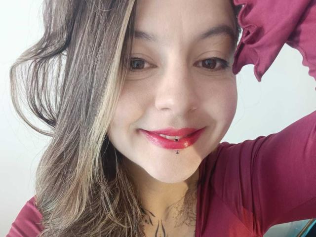 ArianaBlonde with light brown hair, red lipstick, and a lip piercing, wearing a red shirt, smiling at the camera, her right hand in her hair.