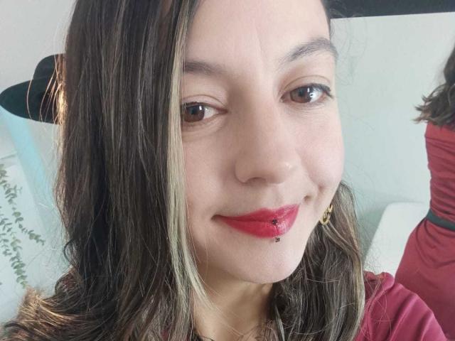 ArianaBlonde has brown eyes, red lipstick, a lip piercing, and dark brown hair with blonde highlights. She's smiling, wearing a red shirt, and has gold earrings.