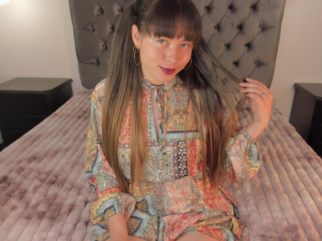 Iniridax ha lunghi capelli castani, indossa un abito colorato con motivi paisley, è seduta su un letto con una testiera grigia imbottita, si tiene i capelli e indossa orecchini a cerchio dorati..