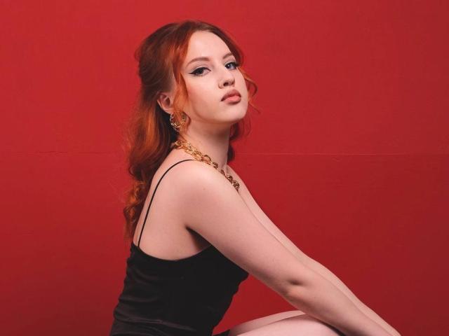 Janna Corsaro, con cabello rojo, vestido negro y collar dorado, sentada frente a un fondo rojo, mostrando sus senos y mirando hacia arriba con una expresión ligeramente fruncida..