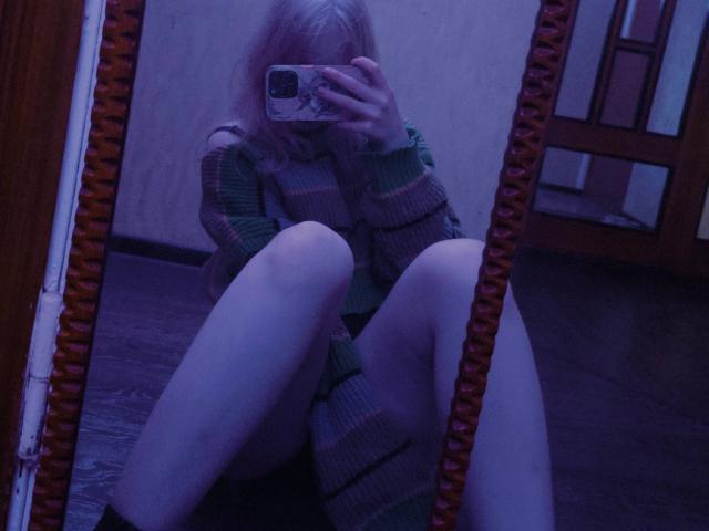 TiffaniMacandog sitting with legs spread, wearing a green striped sweater, taking a mirror selfie with phone, purple-blue lighting, reflection shows her bare ass and knees.
