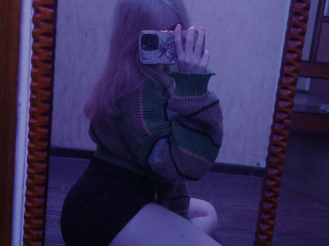 TiffaniMacandog in a green striped sweater, black shorts, kneeling, taking a mirror selfie with phone, showing her ass, purple-tinted reflection, long pinkish hair, wooden mirror frame.
