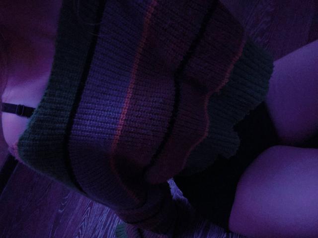 TiffaniMacandog's ass in a green, ribbed, striped sweater, black bra strap visible, sitting on dark wooden floor, purple lighting. Boobs partially visible.