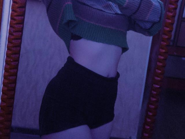 TiffaniMacandog's midriff in a green striped top, black shorts, showing a slim, flat belly with a slight tan line, in a dimly lit room, reflected in a mirror.