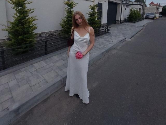 Viloriia in a white dress, holding pink flowers, standing on a street, with her hands in her dress pockets, looking down, blonde hair, small breasts, slim build, daytime.
