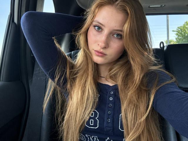 ArdelleCooksey with long blonde hair, blue eyes, and pouty lips, wearing a navy long-sleeve shirt, has her hand behind her head, sitting in a car.