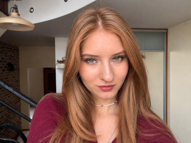 ArdelleCooksey has long, straight, reddish-brown hair, green eyes, and wears a maroon top with a silver choker. Her pink lips are slightly parted, showing a subtle smile.