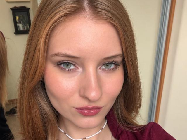 ArdelleCooksey has light green eyes, pink lipstick, and reddish-brown hair, wearing a maroon top and white necklace, with a slightly pouty expression in the photograph.